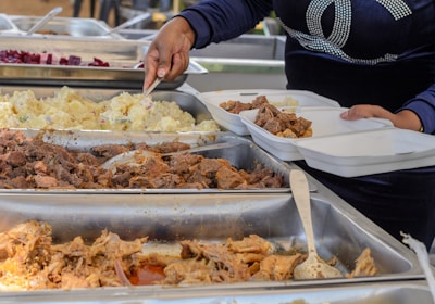 A person is serving food from buffet trays containing various dishes like meat, potatoes, and another food item into divided takeaway containers. The person is using a serving utensil and is wearing a dark blue long-sleeved shirt.